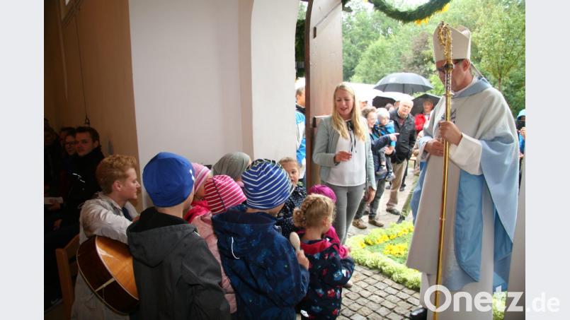 Am Kirchenportal begrüßen die Mädchen und Buben des Gebenbacher Kindergartens mit ihrer Leiterin Gabi Neuberger (links) Weihbischof Josef Graf mit einem Lied. Bild: swo
