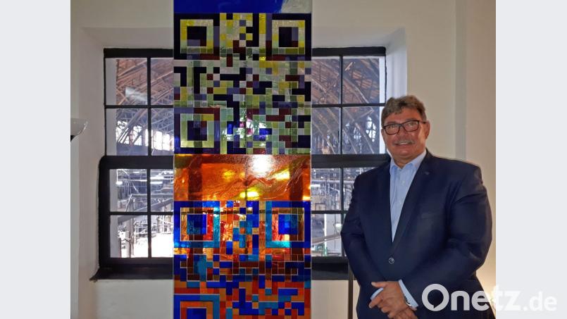 Rainer Schmitt neben einem Glaskunstwerk, das schon vor einigen Jahren bei einer Baumesse präsentiert wurde. Es zeigt die QR-Codes der Glashütte Lamberts und von Schmitts Derix Glasstudios. Es verarbeitet das in handwerklicher Tradition gefertigte Produkt seit vielen Jahren. Bild: pz