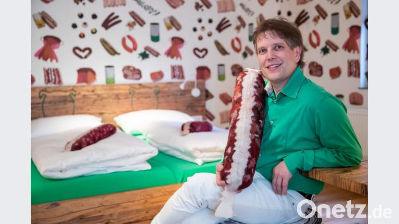Claus Böbel, Metzgermeister und Betreiber des "Bratwursthotels", sitzt mit einem Kissen in Bratwurst-Optik in einem der sieben Zimmer seines Gasthofs. Bild: Daniel Karmann/dpa