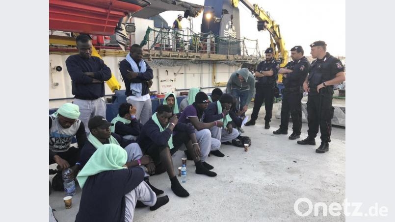 Migranten vom Rettungsschiff &quot;Sea-Watch 3&quot; sitzen bewacht von Polizisten auf dem Steg im Hafen der Insel Lampedusa. Bild: Annalisa Camilli/AP/dpa