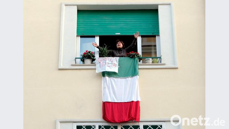 Eine Bewohnerin Roms klatscht und singt am Fenster ihrer Wohnung. An ihrem Fenster hängt eine Landesfahne mit der Aufschrift "Grazie Italia". Bild: Mauro Scrobogna/LaPresse via ZUMA Press/dpa