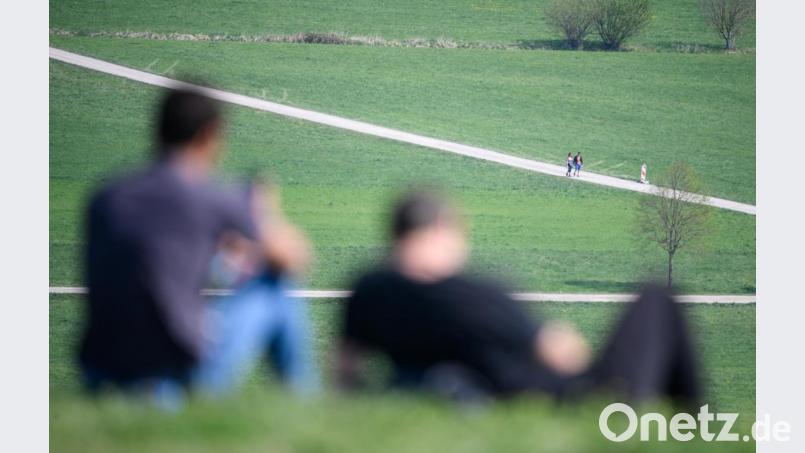 Zwei Männer sitzen auf einem Hügel in der Sonne. Im Hintergrund gehen zwei Spaziergänger. In Thüringen werden die Kontaktbeschränkungen aufgehoben. Bild: Sebastian Gollnow/dpa