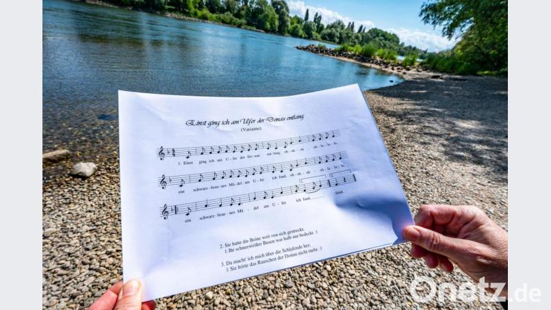 Ein Notenblatt mit dem Liedtext einer Version des Donauliedes wird am Donauufer gehalten. Bild: Armin Weigel/dpa