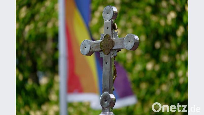Ein Kreuz steht bei einem Open-Air-Segnungsgottesdienst für Liebende vor einer Kirche. Im Hintergrund ist eine Regenbogenfahne zu sehen. Bild: Henning Kaiser/dpa