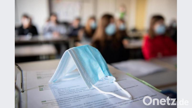 Eine Maske liegt im Unterricht auf Unterlagen, während im Hintergrund Schülerinnen und Schüler in einem Gymnasium mit Mund- und Nasenschutz dem Unterricht folgen. Symbolbild: Matthias Balk