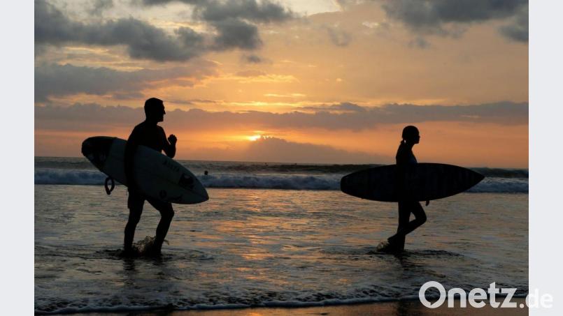 Surfer gehen an einem Strand auf Bali durch seichtes Wasser. Bild: Firdia Lisnawati/AP/dpa/Archivbild
