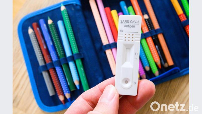 Ein Corona-Selbsttest wird über dem Federmäppchen eines Schülers gehalten. Bild: Julian Stratenschulte/dpa/Illustration