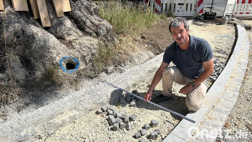 Auch der große Baum in der Kurve der Bahnhofstraße gegenüber der Einfahrt zur Heinrich-Böll-Straße bleibt stehen. Planer Martin Dunst zeigt den dortigen Gehweg, der wie früher in einer Breite von 1,25 Meter daran vorbeiführt. Umgesetzt werden die Vorgaben von der Baufirma Englhard aus Amberg. Bild: Andreas Royer