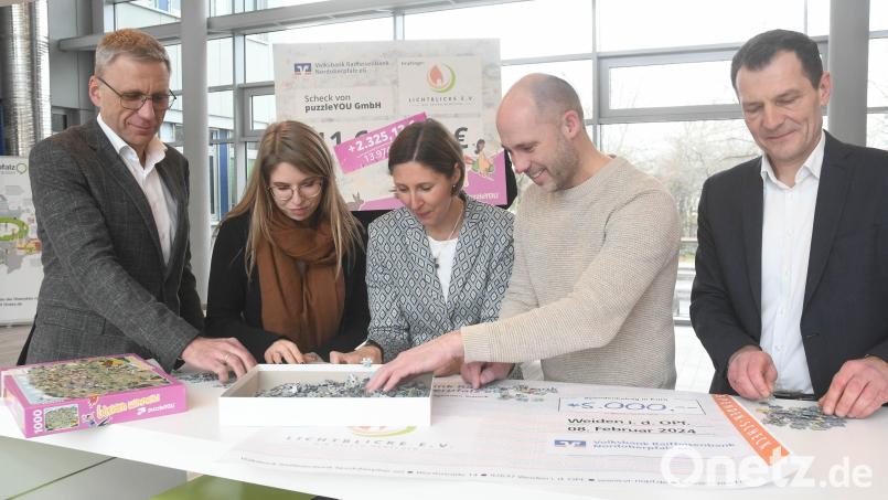 Zwei Schecks – insgesamt über 7000 Euro an Spenden für die Aktion &quot;Lichtblicke.&quot; Bei der Übergabe wird das beliebte Puzzle nochmal ausgepackt. Von links: Bernhard Wolf, Vorstandssprecher der VR-Bank Nordoberpfalz, Nina Lenfers von &quot;puzzleYOU&quot;, Viola Vogelsang-Reichl von &quot;Lichtblicke&quot;, Jens Simon von &quot;puzzleYOU&quot; und Ingo Dietz, Marketing-Manager der VR-Bank Nordoberpfalz. Bild: Gabi Schönberger