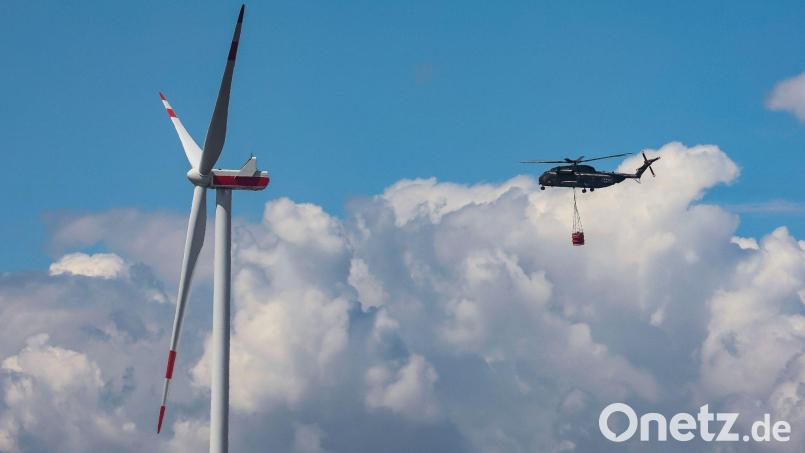 Ein Bundeswehrhubschrauber vom Typ Sikorsky CH-53 fliegt mit Löschwasser zur Waldbrandbekämpfung an einem Windrad vorbei. Bild: Jan Woitas/dpa