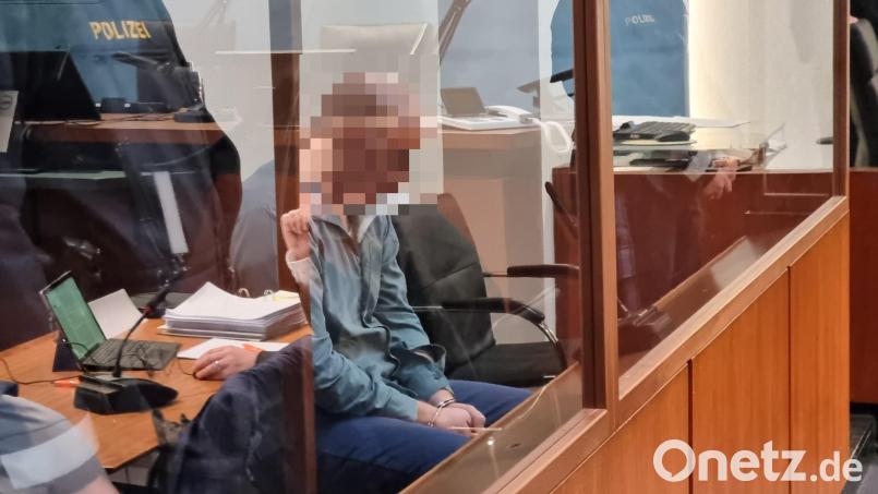 Der Angeklagte sitzt mit gesenktem Kopf auf der Anklagebank hinter einer dicken Glasscheibe. In der Spiegelung ist sein Verteidiger Maximilian Siller erkennbar. Bild: scm