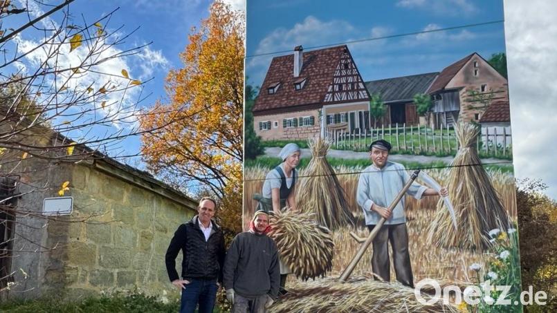 Bürgermeister Hans-Jürgen Strehl mit dem Künstler Markus Ronge vor der Ostseite mit dem Goglhof als Motiv. Wer von Süden kommt sieht die Edelsfelder Windräder, auf der Westseite grüßen die beiden Ortskirchen und im Norden tanzt ein Kirwapaar um den Kirwabaum. Bild: Elfriede Winter