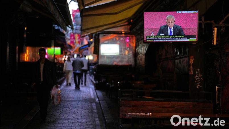 Menschen gehen an einem Fernsehbildschirm vorbei, auf dem der Präsident des Türkischen Fußballverbands, Ibrahim Haciosmanoglu, während einer Pressekonferenz zu sehen ist. Bild: Francisco Seco/AP/dpa