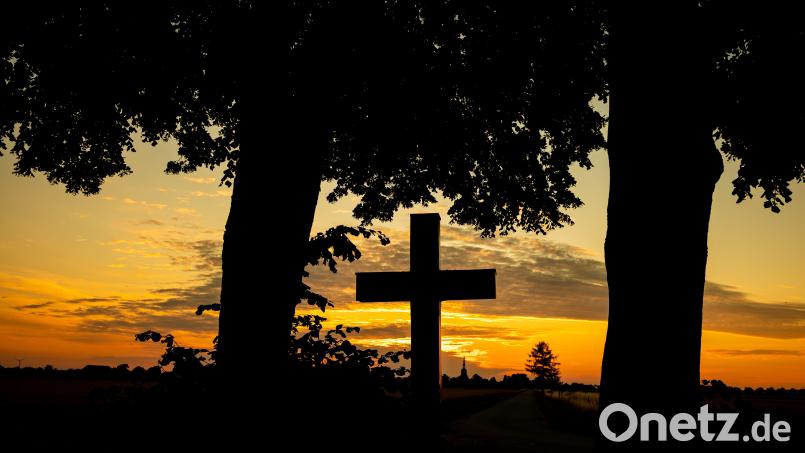 Der Himmel hinter einem Kreuz strahlt kurz vor Sonnenaufgang orange. Friedhöfe sind für viele Angehörige ein besonderer Ort der Trauer. Bild: Moritz Frankenberg/dpa