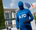 Bild: Paul Zinken/dpa
Alternative Montur: Ein komplett verhüllter Unterstützer der AfD mit der Aufschrift „Verfassungsheld AfD“ auf der Brust seines Ganzkörperanzugs verteilt beim Tag der offenen Tür der Bundesregierung vor dem Kanzleramt Informationsbroschüren der Partei.
