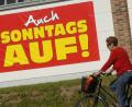 Bild: Stefan Sauer
Eine Radfahrerin fährt an einem großen Plakat mit der Aufschrift "Auch Sonntags auf" vorbei. Foto: Stefan Sauer/Archiv
