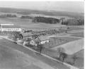 Bild: Bildarchiv der Gemeinde Ursensollen
Diese Luftaufnahme aus dem Jahr 1958 zeigt die damals noch "einsame Lage" des Bahnhofes Ursensollen und der beiden Lagerhäuser südlich des Ortskernes. Westlich der AS 4 findet sich nun das Edeka-Geschäft, südlich zum Beispiel die Firma KTM. Die Abzweigung zur AS 15 in Richtung Hohenkemnath verlief zu dieser Zeit noch mitten durch beide Lagerhausteile. Das Holzlager beim Bahnhof erinnert noch an die West-Erweiterung des Truppenübungsplatzes Hohenfels im Jahre 1951. Damals durften die abgelösten Bauern noch einige Monate in ihren Wäldern einen Holzabschlag durchführen, der Holzhändler aus ganz Deutschland anlockte. Im Hintergrund ist die ehemalige Halle der Firma Ehebauer zu sehen. Später war hier die Firma Schmieding. Nun - seit 2008 - ist hier die Firma "Dr. Dr. Eberth, Arzneimittel" angesiedelt.