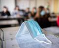 Symbolbild: Matthias Balk
Eine Maske liegt im Unterricht auf Unterlagen, während im Hintergrund Schülerinnen und Schüler in einem Gymnasium mit Mund- und Nasenschutz dem Unterricht folgen.