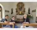 Bild: Veronika Kreuzer
Rita Kreuzer in der Kapelle, um die sie sich schon seit vielen Jahren kümmert. Mit den Enkeln Konstantin und Johann (von links) ist schon die nächste Generation bereit. Am Altar stehen die Figuren des heiligen Michael (links) und der heiligen Margareta (rechts). An den Wänden sind auf einem Sockel Statuen des heiligen Antonius (links) und der Muttergottes (rechts) angebracht.