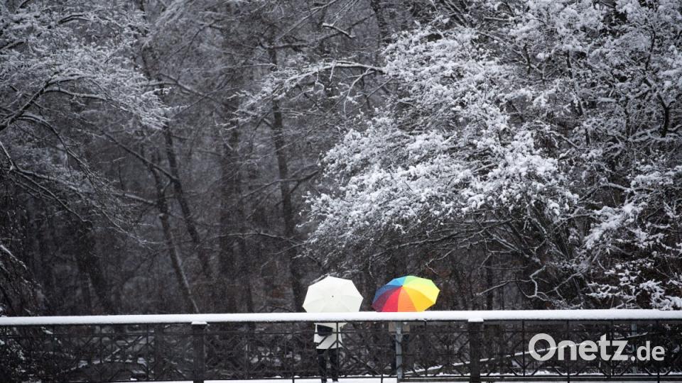 Ein Paar mit Regenschirmen steht im Nymphenburger Schlossgarten auf einer schneebedeckten Brücke. Bild: Lino Mirgeler/dpa