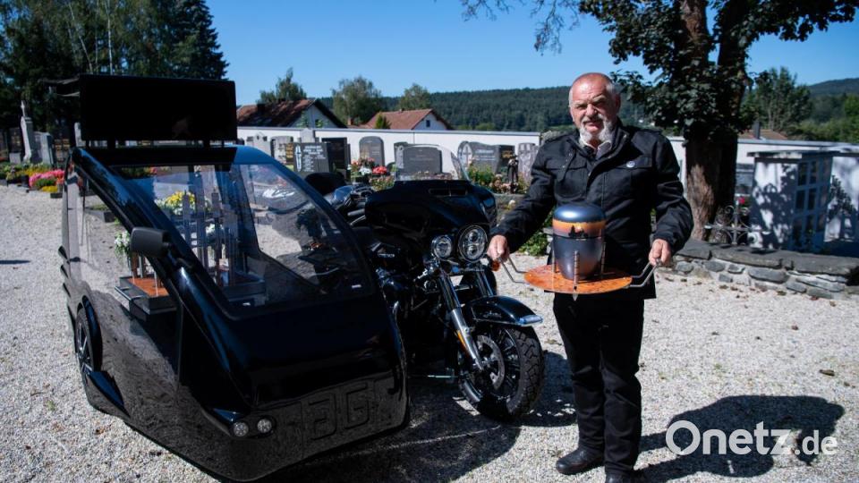 Bestatter Wolfgang Frisch steht mit seinem Bestattungsmotorrad, einer umgebauten Harley-Davidson, und einer Urne auf einen Friedhof. Bild: Sven Hoppe/dpa