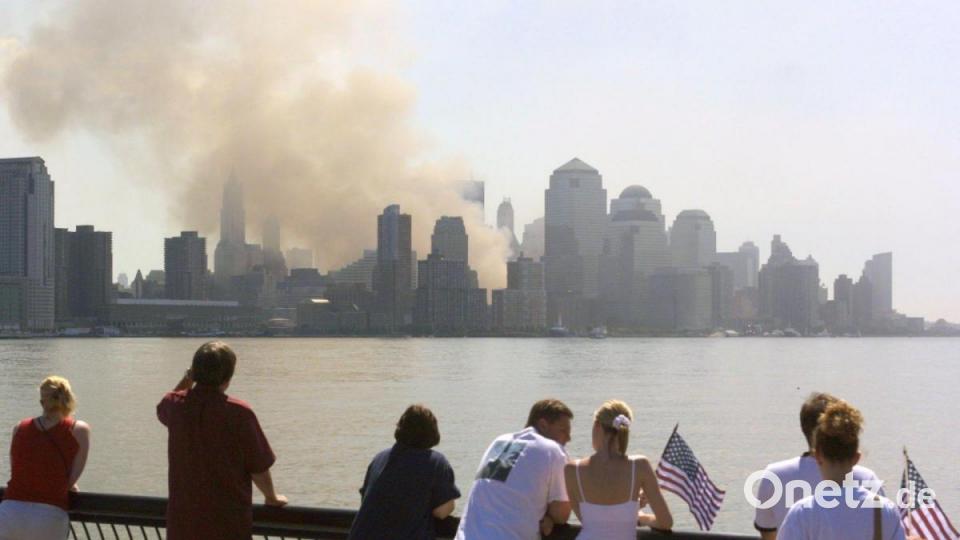 Passanten mit US-Fahnen betrachten von New Jersey aus die in Rauch und Staub gehüllte Skyline von Manhattan. Dort wo am Vortag noch die beiden Türme des World Trade Centers gestanden hatten, ist jetzt nur noch eine Qualmwolke zu sehen. Attentäter hatten am Vortag insgesamt vier Passagiermaschinen entführt. Zwei lenkten sie dabei auf die beiden Türme des World Trade Center, eins auf das Washingtoner Pentagon. Ein viertes stürzte bei Pittsburgh ab. Archivbild: Hubert Boesl/dpa