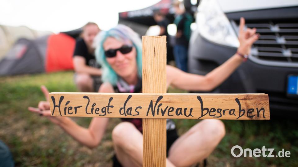 Eine Frau sitzt auf dem Wacken Open Air Festival hinter einem Holzkreuz mit der Aufschrift „"Hier liegt das Niveau begraben".“ Bild: Daniel Reinhardt/dpa