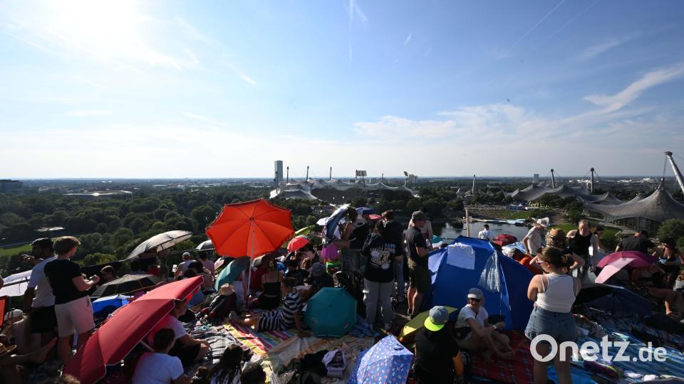 Fans sitzen mit Sonnenschirmen vor der Kulisse des Olympiastadions auf dem Gipfel des Olympiabergs. Bild: Felix Hörhager /dpa
