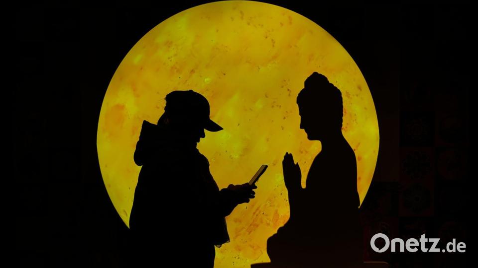 "Zwiegespräch": Eine Frau surft auf ihrem Smartphone vor einer Buddha-förmigen Pappfigur, die in einem Einkaufszentrum in Peking aufgestellt ist. Bild: Andy Wong/AP/dpa