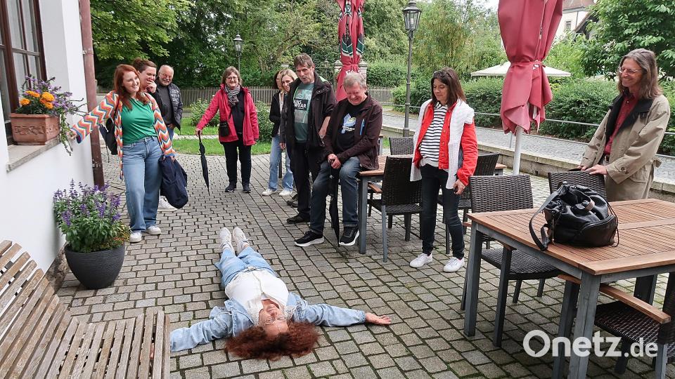 Zur Freude der Teilnehmer wurden bei der Drehort-Tour durch Friedenfels auch Szenen nachgespielt, wie hier von Ulla Britta Baumer (auf dem Boden liegend) die Szenerie, als sie im Film mausetot vor der Eingangstür des Friedenfelser Dorfladens liegen musste. Bild: bsc