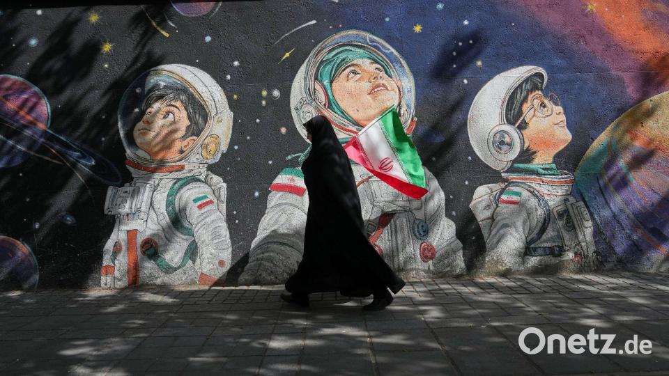 Mit einer iranischen Flagge in der Hand geht eine Frau an einer Wand vorbei, auf der Kinder in Raumanzügen zu sehen sind. Bild: Francisco Seco/AP/dpa
