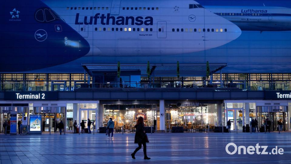 Passagiere gehen am Münchner Flughafen vor dem Terminal 2 an einem Plakat mit Flugzeugen der Lufthansa entlang. Bild: Sven Hoppe/dpa
