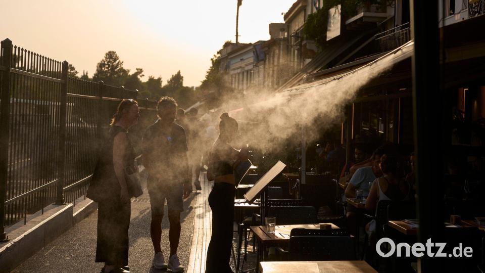 Ein Touristenpaar geht an einer Sprühanlage vorbei, während ein Restaurantmitarbeiter sie im Athener Stadtteil Monastiraki zu ihrem Tisch führt. Bild: Petros Giannakouris/AP/dpa