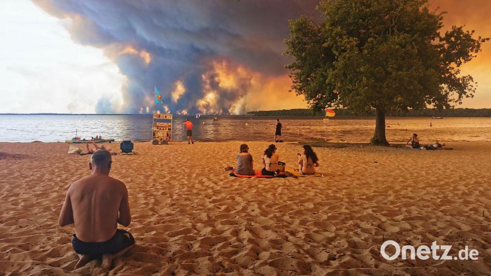 Strandbesucher sitzen am Strand von Moutchic in Lacanau im Südwesten Frankreichs, während Rauchwolken von einem Waldbrand in den Himmel aufsteigen. Bild: Maximilien Lamy/AFP/dpa