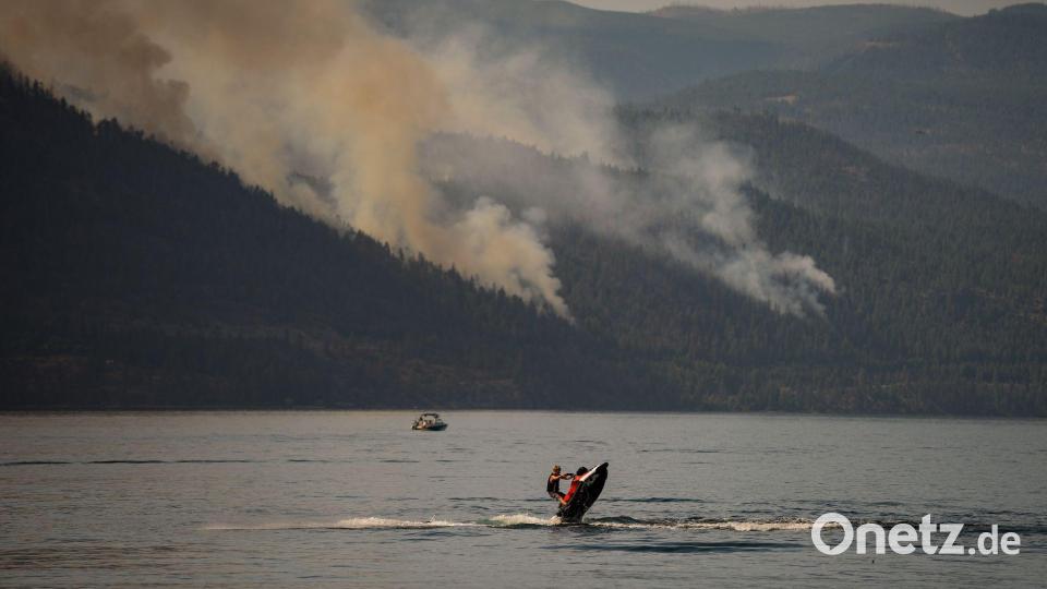 Eine Person fährt mit einem Jetski in Kanada, währen im Hintergrund ein Wald brennt. Bild: DARRYL DYCK/The Canadian Press/dpa