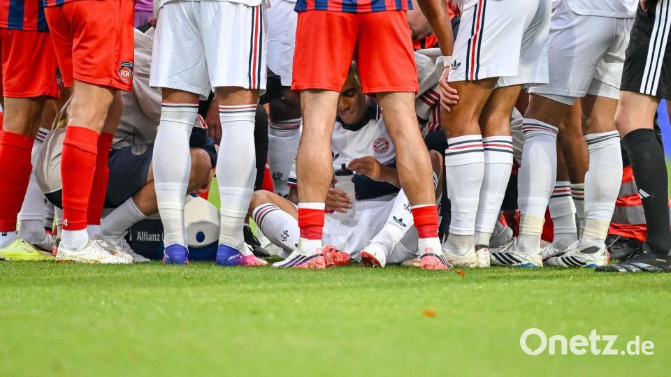Jamal Musiala sitzt, abgeschirmt von Münchner Teamkollegen und Heidenheimer Fußballern, nach seinem Zusammenbruch auf dem Rasen. Bild: Harry Langer/DeFodi/dpa/dpa
