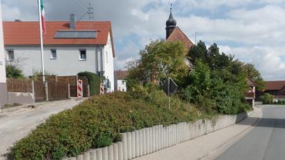 Bild: Brendel, Hubert (BR)
Die Betonpalisaden südlich der Sankt Anna Kirche (Mitte, im Hintergrund) sollen bei den Maßnahmen im Zuge der einfachen Dorferneuerung, vorbehaltlich einer Förderung, durch eine dorfgerechte Trockenmauer aus Granitsteinen ersetzt werden. Gleichzeitig soll das schadhafte Betonpflaster im Gehweg erneuert werden. Die dafür veranschlagten Kosten belaufen sich auf rund 54 000 Euro.