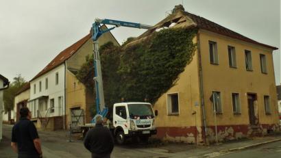 Bild: jml
Der Abriss beginnt. Von oben nach unten wird das alte Beer-Walbrunn-Geburtshaus entblättert. Die Kunstschiefer-Dachdeckung muss als Sondermüll behandelt werden und bei der imposanten Efeupflanze ist entblättern wörtlich zu nehmen. Karlheinz Meier, stellvertretender Vorsitzender des Kunst- und Kulturvereins Anton Beer- Walbrunn und ein großer Befürworter des Erhalts des Gebäudes (rechts), beobachtet das Geschehen.
