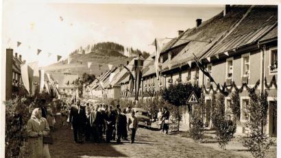 Bild: hl
Dieses historische Bild zeigt eine Fronleichnamsprozession durch Waldeck in den 1950er Jahren. Vor der Blaskapelle gingen damals viele Schulkinder, begleitet von einem Lehrer. Fast jedes Haus hatte eine blau-weiße Fahne aufgezogen. Erst gegen Mitte des Jahrzehnts mischten sich darunter Fahnen mit den Marktfarben rot-gelb-grün. An den Häusern waren Girlanden aus Fichtenstreu angebracht und an den Fenstern im ersten Stock hängten die Bewohner rote Tücher auf.
