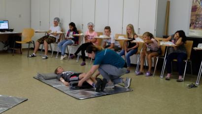 Bild: exb/Bayerisches Rotes Kreuz Kreisverband Tirschenreuth
Natalie Wölfl vom Jugendrotkreuz zeigt den Fünftklässlern der Otto-Wels-Mittelschule Mitterteich, wie bei Bauchschmerzen mit der „Knierolle“ der Patienten in eine Schonhaltung gebracht werden kann.