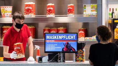 Bild: Robert Michael/dpa
Auf einem Bildschirm im Foyer des Kinos Ufa-Kristallpalast in der Innnenstadt von Dresden wird auf die Maskenpflicht hingewiesen, während ein Mitarbeiter an der Theke Popcorn verkauft.