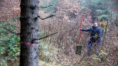 Bild: bkr
Eingezäunt mitten im Wald ist eine der fünf zu sanierenden Trinkwasserquellen. Bürgermeister Thomas Kaufmann steht am Quellstein. Nur dieser ist sichtbar. Die Quellfassung selbst liegt im Verborgenen und muss zur Sanierung sensibel geöffnet werden. Der rote Ring am Baum bedeutet, dass er gefällt werden muss.