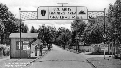 Archivbild: Fotohaus Spahn/exb
Bis Juli 1990 stand das alte Holzhäuschen am Lagereingang, des berühmten „Gate 1“. Das Motiv mit der jungen Frau, zwei Soldaten der Wachkompanie und dem markanten Torbogen wurde in den 1950er und 60er Jahren auf Tausenden von Postkarten und Bildern in der ganzen Welt verbreitet.