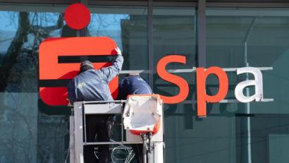 Bild: Sebastian Kahnert/dpa
Die Sparkassen kommen bisher besser durch die Corona-Krise als befürchtet: Zwei Werbetechniker installieren mit einer Hebebühne ein Logo der Sparkasse an einer Glasfassade.