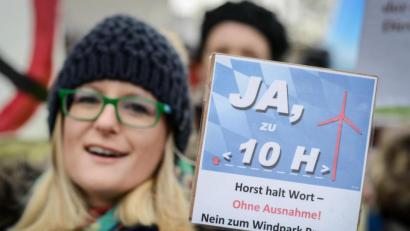 Bild: David Ebener/dpa
Einer Legende zufolge soll Horst Seehofer auf die 10-H-Regelung gekommen sein, als er mit seinem Dienstwagen an einer Gruppe Windkraftgegner vorbeifuhr, seinen Chauffeur halten ließ und mit den Demonstranten diskutierte. Im Bild: Eine Gegnerin des Ausbaus der Windenergie in Bayern demonstriert am 15. Februar 2014 vor Beginn des kleinen Parteitages der CSU in Bamberg (Bayern) mit einem Schild mit der Aufschrift "Ja zu 10 H - Horst halt Wort - ohne Ausnahme ! Nein zum Windpark Brunn!!!".