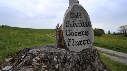 Symbolbild: Karl-Josef Hildenbrand/dpa
An einem auf einem Baumstumpf stehenden Kreuz lehnt eine Holzscheibe mit der Aufschrift "Gott schütze unsere Fluren".