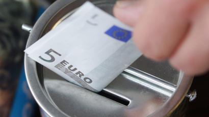 Symbolbild: Bernd Thissen/dpa
Ein Fünf-Euro-Schein wird in eine Spendendose gesteckt.