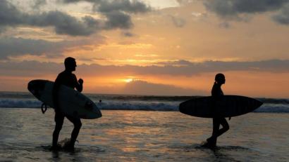Bild: Firdia Lisnawati/AP/dpa/Archivbild
Surfer gehen an einem Strand auf Bali durch seichtes Wasser.