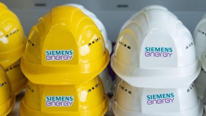 Bild: Sebastian Kahnert/dpa-Zentralbild/dpa/Symbolbild
Schutzhelme mit der Aufschrift „Siemens Energy“ liegen in einer Halle auf einem Tisch.