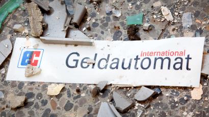 Symbolbild: Patrick Pleul
Splitter und ein Schild mit der Aufschrift "Geldautomat" liegen auf dem Fußboden vor einem gesprengten Geldautomaten.